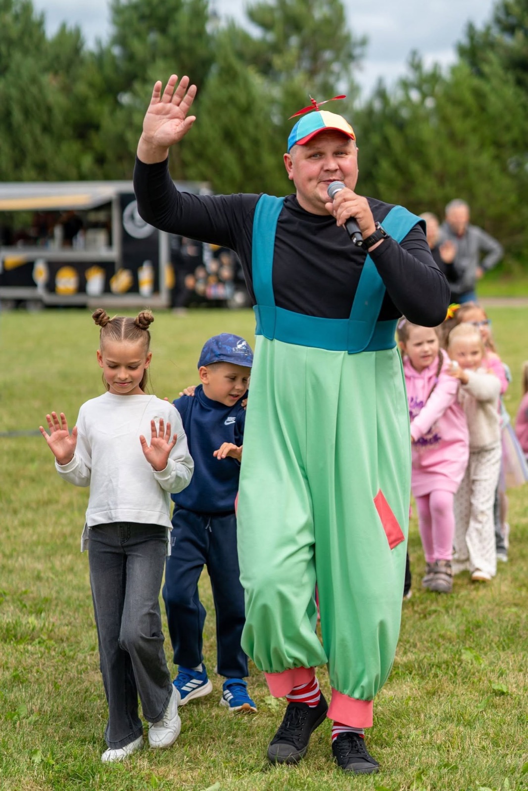 Atlikėjas su sraigtine kepuraite veda vaikų traukinuką per pievą